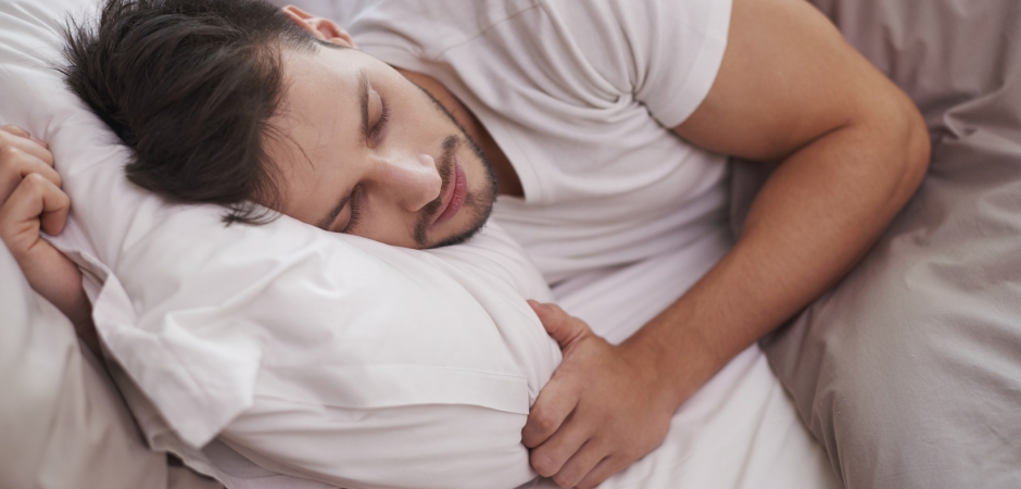The image depicts a man in a state of deep sleep. He rests comfortably on his back, his head nestled on a soft pillow. The gentle rise and fall of his chest indicates his calm, steady breathing. This simple scene conveys the importance of a good night's sleep for overall health and well-being. The pillow, a key element, provides proper support for the neck and head, promoting a restful sleep posture.